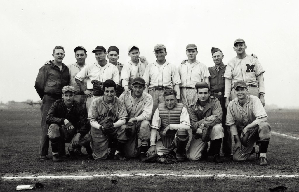 United States Army Air Force ballplayers. There is a hodgepodge of uniforms on display in this image. In addition to the Cincinnati and "M" jerseys, the player in the front row (2nd from right) is wearing a uniform with thin, double-piping). The player in the bomber jacket (front row, far left) is wearing a 6 panel hat with piping outlining each panel.